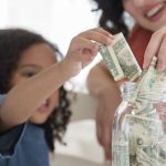 A young girl adds dollar bills to a savings jar, while an adult supports her money-saving activity in a bright home setting.