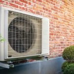 Air conditioning unit on a textured brick wall with trimmed shrubs, showcasing outdoor climate control functionality.