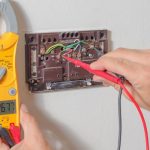 A person uses a Fieldpiece SC260 clamp meter to test the voltage of a thermostat's wiring. The meter reads 26.77 volts.