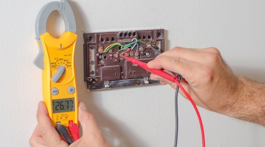 A person uses a Fieldpiece SC260 clamp meter to test the voltage of a thermostat's wiring. The meter reads 26.77 volts.
