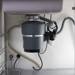 Gray garbage disposal unit under kitchen sink with visible pipes and electrical connections for home maintenance.